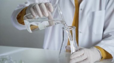 Laboratuvar önlüğü ve eldiven giyen bilinmeyen bilim adamı dikkatlice bir şişeden bir sıvıyı Erlenmeyer şişesine boşaltıyor. Bilim ve tıp.