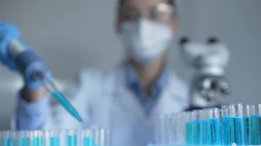 Laboratuvar önlüğü, maskesi ve güvenlik gözlüğü takan kadın araştırmacı test tüpüne mavi bir sıvı atmak için mikropipet kullanıyor. Tıp ve bilim konsepti.