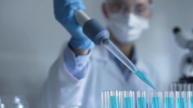 Laboratuvar önlüğü, maskesi ve güvenlik gözlüğü takan kadın araştırmacı test tüpüne mavi bir sıvı atmak için mikropipet kullanıyor. Tıp ve bilim konsepti.