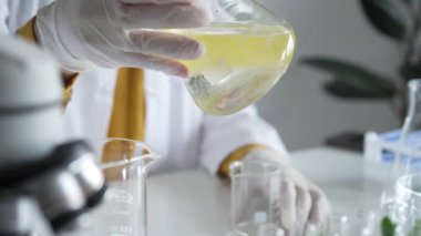 Beyaz koruyucu eldiven giyen bilinmeyen bilim adamı, bir laboratuarda, bir Erlenmeyer şişesinin içinde sarı bir kimyasal çözeltiyi dikkatlice sallıyor. Bilim ve tıp.