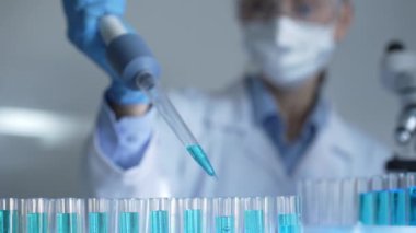 Laboratuvar önlüğü, maskesi ve güvenlik gözlüğü takan kadın araştırmacı test tüpüne mavi bir sıvı atmak için mikropipet kullanıyor. Tıp ve bilim konsepti.