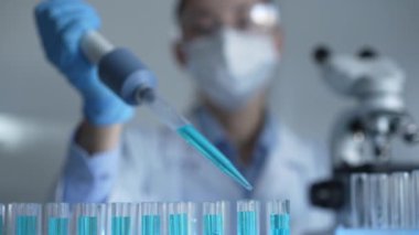 Laboratuvar önlüğü, maskesi ve güvenlik gözlüğü takan kadın araştırmacı test tüpüne mavi bir sıvı atmak için mikropipet kullanıyor. Tıp ve bilim konsepti.