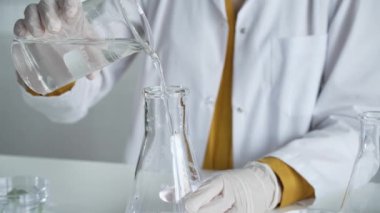 Laboratuvar önlüğü ve eldiven giyen bilinmeyen bilim adamı dikkatlice bir şişeden bir sıvıyı Erlenmeyer şişesine boşaltıyor. Bilim ve tıp.