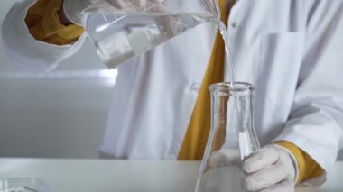 Laboratuvar önlüğü ve eldiven giyen bilinmeyen bilim adamı dikkatlice bir şişeden bir sıvıyı Erlenmeyer şişesine boşaltıyor. Bilim ve tıp.