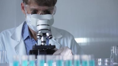 Laboratuvar önlüğü, maske, beyaz eldivenler ve güvenlik gözlüğü takan erkek bilim adamı laboratuarda test tüpleriyle çevrili bir dürbün kullanıyor. Tıp ve bilim konsepti.