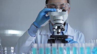 Laboratuvar önlüğü, maske, mavi eldiven ve güvenlik gözlüğü takan bilim adamı kadın, laboratuarda test tüpleriyle çevrili bir mikroskobu ayarlıyor. Tıp ve bilim konsepti.
