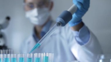 Laboratuvar önlüğü, maskesi ve güvenlik gözlüğü takan kadın araştırmacı test tüpüne mavi bir sıvı atmak için mikropipet kullanıyor. Tıp ve bilim konsepti.