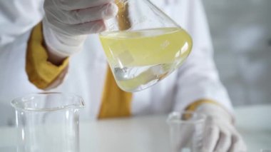 Beyaz koruyucu eldiven giyen bilinmeyen bilim adamı, bir laboratuarda, bir Erlenmeyer şişesinin içinde sarı bir kimyasal çözeltiyi dikkatlice sallıyor. Bilim ve tıp.