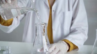 Laboratuvar önlüğü ve eldiven giyen bilinmeyen bilim adamı dikkatlice bir şişeden bir sıvıyı Erlenmeyer şişesine boşaltıyor. Bilim ve tıp.