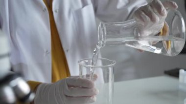 Laboratuvar önlüğü ve eldiven giyen bilinmeyen bilim adamı laboratuvar setinde bir şişeden diğerine sıvıyı dikkatlice döküyor, kapat. Bilim ve tıp kavramları.