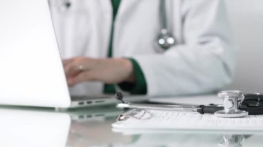 Stetoskop ve ilaç geçmişi kayıtları, klinik ofis arka planında dizüstü bilgisayarla çalışan bir doktorun önünde cam bir masanın üzerinde yatıyor. Tıp konsepti.