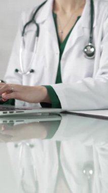 Doktor önlüğünün altında yeşil bluz giyen bir kadın doktor sağlık hizmetlerinde teknoloji kullanıyor. Klinikteki cam bir masanın üstünde dizüstü bilgisayara dikey bir bakış açısı olan ellerin yakın çekimi. Tıbbi konsept.