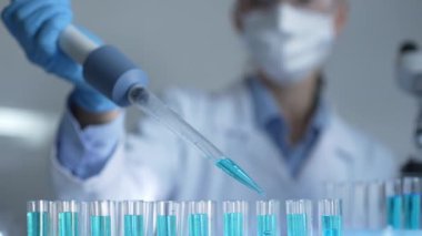 Laboratuvar önlüğü, maskesi ve güvenlik gözlüğü takan kadın araştırmacı test tüpüne mavi bir sıvı atmak için mikropipet kullanıyor. Tıp ve bilim konsepti.