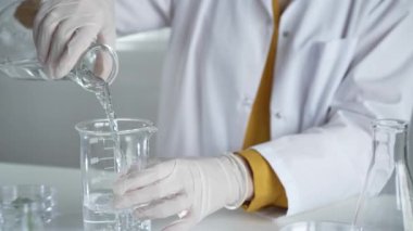 Laboratuvar önlüğü ve eldiven giyen bilinmeyen bilim adamı laboratuvar setinde bir şişeden diğerine sıvıyı dikkatlice döküyor, kapat. Bilim ve tıp kavramları.