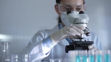 Laboratuvar önlüğü, maskesi, beyaz eldivenleri ve güvenlik gözlüğü takan bilim adamı bir kadın laboratuarda test tüpleriyle çevrili bir mikroskop kullanıyor. Portre görüntüsü. Tıp ve bilim konsepti.