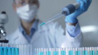 Laboratuvar önlüğü, maskesi ve güvenlik gözlüğü takan kadın araştırmacı test tüpüne mavi bir sıvı atmak için mikropipet kullanıyor. Tıp ve bilim konsepti.