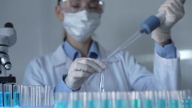 Laboratuvar önlüğü, maske ve güvenlik gözlüğü takan bilim adamı kadın araştırmacı test tüplerine sıvı atmak için bir mikropipet kullanıyor. Tıp ve bilim konsepti.