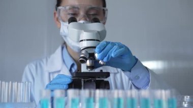 Laboratuvar önlüğü, maske, mavi eldiven ve güvenlik gözlüğü takan bilim adamı kadın, laboratuarda test tüpleriyle çevrili bir mikroskobu ayarlıyor. Tıp ve bilim konsepti.
