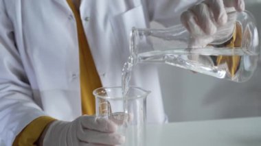 Laboratuvar önlüğü ve eldiven giyen bilinmeyen bilim adamı laboratuvar setinde bir şişeden diğerine sıvıyı dikkatlice döküyor, kapat. Bilim ve tıp kavramları.