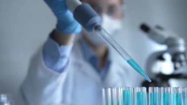 Laboratuvar önlüğü, maskesi ve güvenlik gözlüğü takan kadın araştırmacı test tüpüne mavi bir sıvı atmak için mikropipet kullanıyor. Tıp ve bilim konsepti.