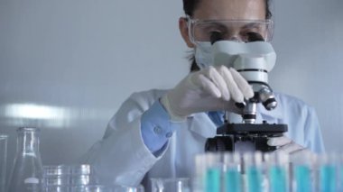 Laboratuvar önlüğü, maskesi, beyaz eldivenleri ve güvenlik gözlüğü takan bilim adamı bir kadın laboratuarda test tüpleriyle çevrili bir mikroskop kullanıyor. Portre görüntüsü. Tıp ve bilim konsepti.