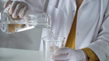 Laboratuvar önlüğü ve eldiven giyen bilinmeyen bilim adamı laboratuvar setinde bir şişeden diğerine sıvıyı dikkatlice döküyor, kapat. Bilim ve tıp kavramları.