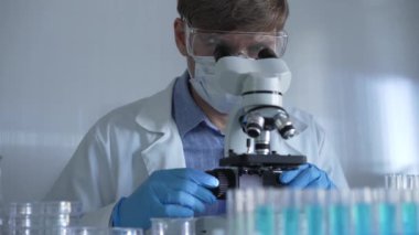 Laboratuvar önlüğü, maske, mavi eldiven ve güvenlik gözlüğü takan bilim adamı laboratuarda test tüpleriyle çevrili bir dürbün kullanıyor. Portre görüntüsü. Tıp ve bilim konsepti.