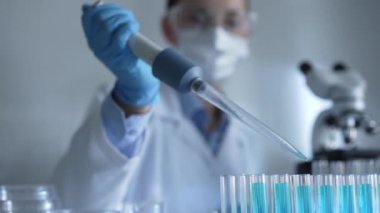 Laboratuvar önlüğü, maskesi ve güvenlik gözlüğü takan kadın araştırmacı test tüpüne mavi bir sıvı atmak için mikropipet kullanıyor. Tıp ve bilim konsepti.