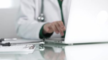 Stetoskop ve ilaç geçmişi kayıtları, klinik ofis arka planında dizüstü bilgisayarla çalışan bir doktorun önünde cam bir masanın üzerinde yatıyor. Tıp konsepti.