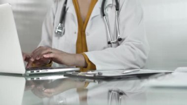 Tıbbi önlüğün altına sarı bluz giyen kadın doktor, sağlık hizmetlerinde teknolojiyi kullanıyor, bilgisayarında stetoskop, gözlük ve not panosuyla yazı yazan elleri kapalı. Tıp konsepti.