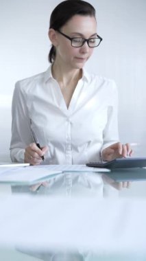 Beyaz günlük bluz ve gözlük giyen iş kadını veya muhasebeci, vergi hesaplamaları, not alma ve ofisteki mali raporlar üzerinde çalışıyor. Ticarette denetim ve vergiler.