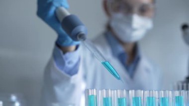 Laboratuvar önlüğü, maskesi ve güvenlik gözlüğü takan kadın araştırmacı test tüpüne mavi bir sıvı atmak için mikropipet kullanıyor. Tıp ve bilim konsepti.