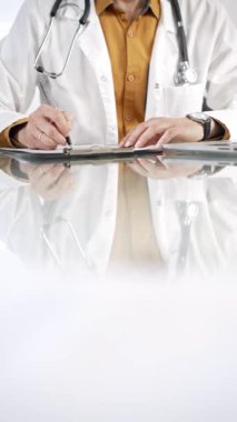 Stetoskop giyen, laboratuvar önlüğünün altına sarı tişört giyen kimliği belirsiz bir erkek doktor, pano kullanıyor ve tıp bürosundaki cam masaya notlar alıyor. Tıp konsepti.