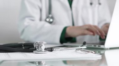 Stetoskop ve ilaç geçmişi kayıtları, klinik ofis arka planında dizüstü bilgisayarla çalışan bir doktorun önünde cam bir masanın üzerinde yatıyor. Tıp konsepti.