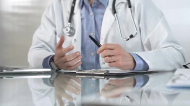 Mavi gömlek, steteskop ve laboratuvar önlüğü giyen erkek doktor, cam bir masadaki hastaya dizüstü bilgisayarla teşhisi açıklarken elleriyle hareket ediyor. Tıp konsepti.