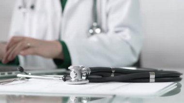 Stetoskop ve ilaç geçmişi kayıtları, klinik ofis arka planında dizüstü bilgisayarla çalışan bir doktorun önünde cam bir masanın üzerinde yatıyor. Tıp konsepti.