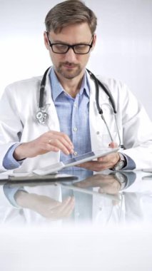 Beyaz önlük, gözlük ve steteskop altında mavi gömlekli doktor modern sağlık ortamında dijital tablet kullanıyor. Hasta verileriyle, dikey portre görüntüsüyle etkileşimde. Tıp ve Bilim.