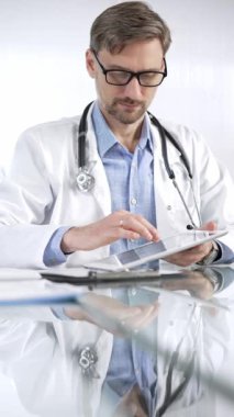 Beyaz önlük, gözlük ve steteskop altında mavi gömlekli doktor modern sağlık ortamında dijital tablet kullanıyor. Hasta verileriyle, dikey portre görüntüsüyle etkileşimde. Tıp ve Bilim.