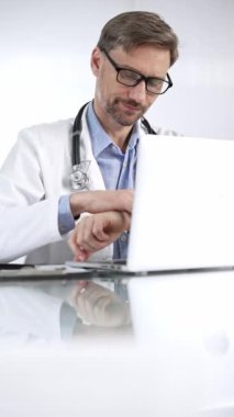 Mavi gömlekli doktor tıp önlüğünün altında teknolojiyi kullanıyor. Klinikteki cam bir masada dizüstü bilgisayarda dikey bir şekilde yazı yazan bir doktorun portresi. Tıp ve Bilim.