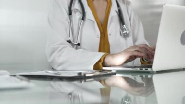 Tıbbi önlüğün altına sarı bluz giyen kadın doktor, sağlık hizmetlerinde teknolojiyi kullanıyor, bilgisayarında stetoskop, gözlük ve not panosuyla yazı yazan elleri kapalı. Tıp konsepti.