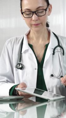 Modern sağlık ortamında dijital tablet kullanan beyaz önlüklü kadın doktor hasta verileriyle ve dikey portreyle etkileşimde. Tıp konsepti.