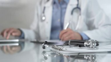Stetoskop ve ilaç geçmişi kayıtları cam bir masanın üzerinde duruyor. Klinik ofis arka planında dizüstü bilgisayarla çalışan bir erkek doktorun önünde. Tıp konsepti.