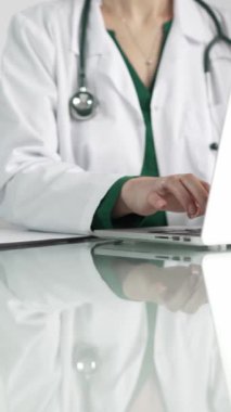 Doktor önlüğünün altında yeşil bluz giyen bir kadın doktor sağlık hizmetlerinde teknoloji kullanıyor. Klinikteki cam bir masanın üstünde dizüstü bilgisayara dikey bir bakış açısı olan ellerin yakın çekimi. Tıbbi konsept.
