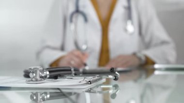 Stetoskop ve ilaç geçmişi kayıtları cam bir masanın üzerinde sarı bluzlu kadın bir doktorun önünde beyaz bir sağlık önlüğünün altında duruyor. Doktor klinik ofis geçmişi üzerinde çalışıyor. İlaç.