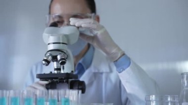 Laboratuvar önlüğü, maskesi, beyaz eldivenleri ve güvenlik gözlüğü takan bilim adamı bir kadın laboratuarda test tüpleriyle çevrili bir mikroskop kullanıyor. Portre görüntüsü. Tıp ve bilim konsepti.