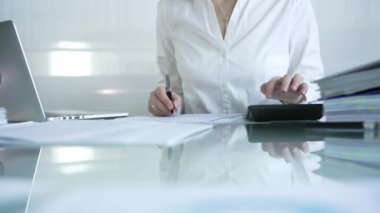 Mavi bluzlu iş kadını, çağdaş ofis ortamında finansal rapor üzerinde çalışırken siyah hesap makinesi kullanıyor ve not alıyor. Denetim ve vergiler.