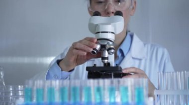 Laboratuvar önlüğü ve güvenlik gözlüğü takan bilim adamı bir kadın laboratuarda test tüpleriyle çevrili bir mikroskop kullanıyor. Tıp ve bilim konsepti.