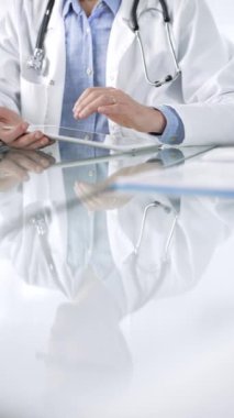 Modern sağlık ortamında dijital tablet kullanan, hasta verileriyle etkileşim kuran, dikey görüşlü, mavi gömlekli, beyaz önlüklü tanınmayan bir doktor. İlaç ve sağlık hizmetleri kavramı.