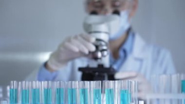 Mavi bir sıvıyla dolu laboratuvar tüpleri odaklanmış, yakından bakıyor. Kadın araştırmacı, arka planda mikroskop kullanıyor. Tıp ve bilim konsepti.