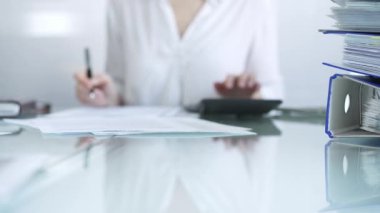 Finansal belgelerin klasörleri cam masanın üzerinde duruyor ve hesap makinesiyle kadın muhasebecinin yanında işlem için bekliyor ve ofiste notlar alıyor. Vergiler, iş konsepti denetimi.